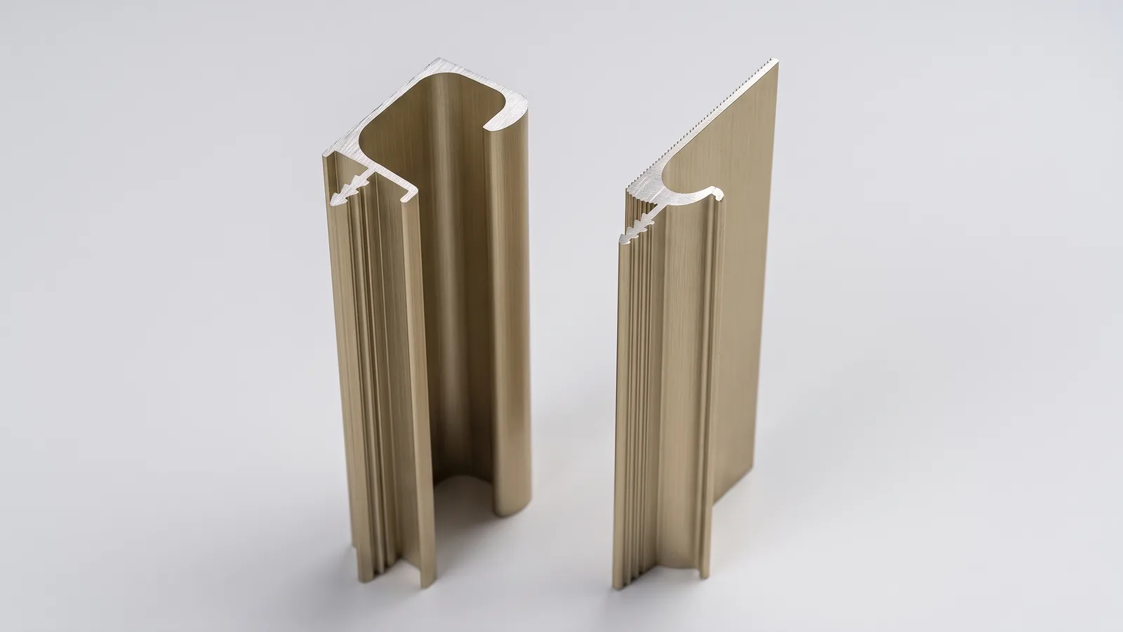 Cross-section ends of a champagne gold G-section profile and an Eureka J profile standing upright on a light grey backdrop